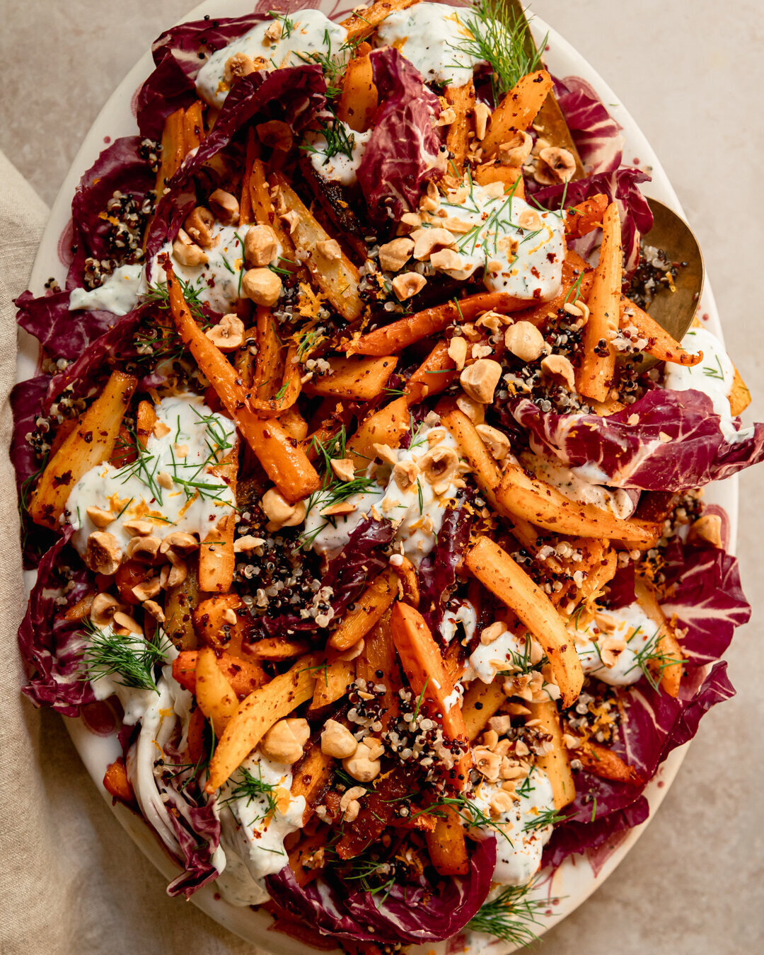 An up close, overhead shot of a harissa carrot and parsnip salad with cooked quinoa, chopped fresh dill, a creamy vegan yogurt sauce dolloped on top, and chopped toasted hazelnuts. All of this is set on a bed of radicchio leaves in a platter