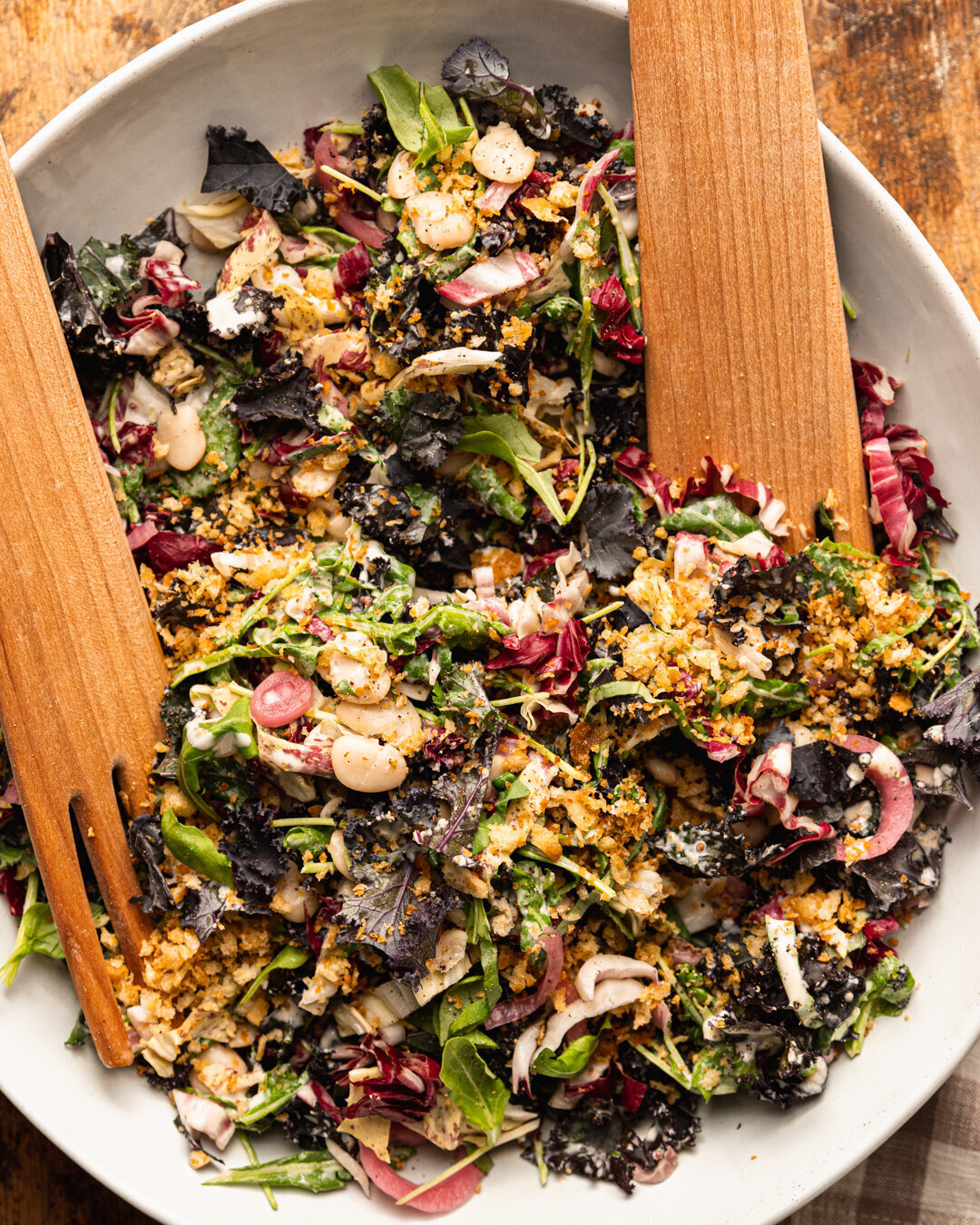 An overhead shot of a kale, arugula and butter bean salad with pickled red onions and crunchy garlicky bread crumbs on top. The salad is in a large, organically shaped bowl with wooden serving tons sticking out.