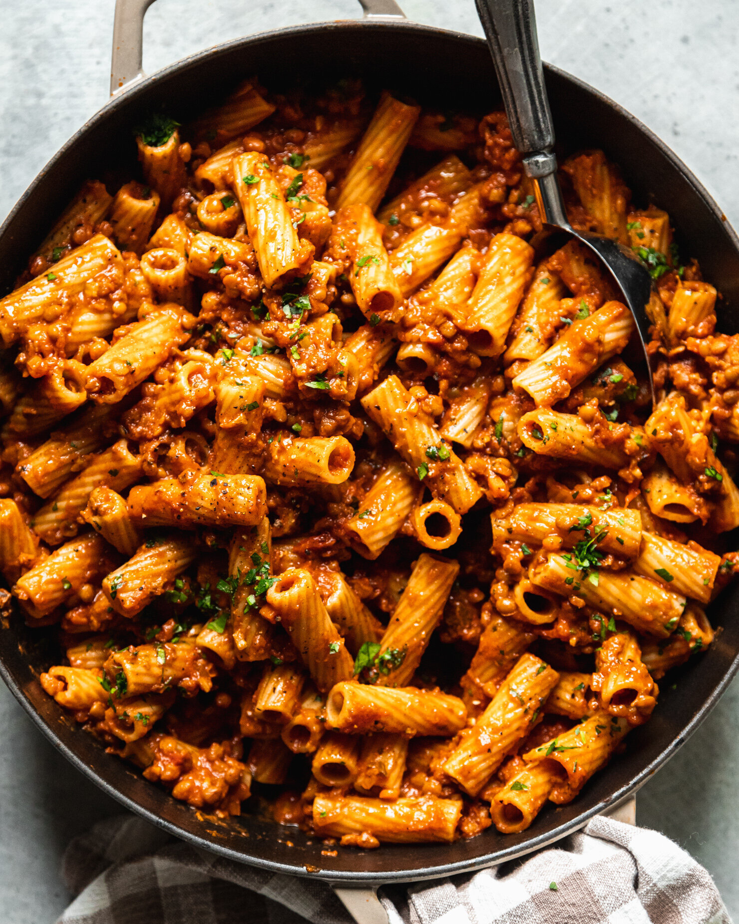 An up close, overhead shot shows rigatoni pasta in a braiser-style pot that's just covered in a thick and meaty lentil bolognese sauce. The finished pasta is garnished with finely chopped parsley.
