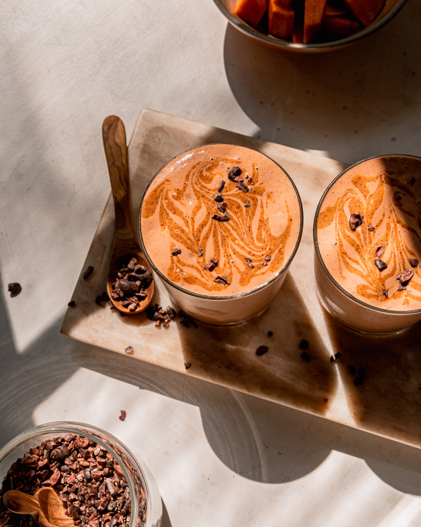 An overhead shot shows two chocolate sweet poytato smoothies garnished with almond butter swirls and cacao nibs. The smoothies are on a marble tray and shown in direct sunlight.