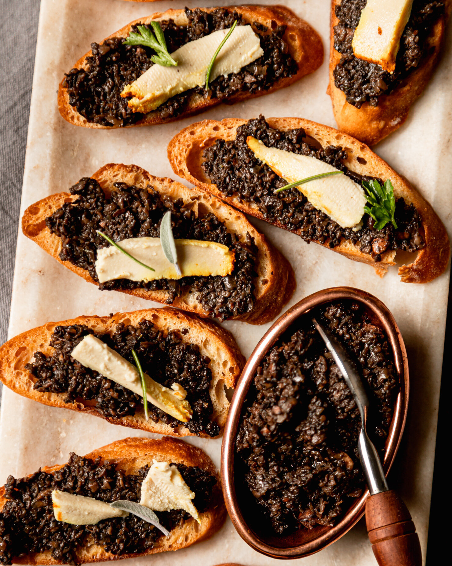 An overhead shot shows crostinis topped with mushroom duxlles and little slivers of soft vegan cheese. Whole leaf herbs are used as garnish and a small bowl of the duxelles is nearby with a spoon sticking out.