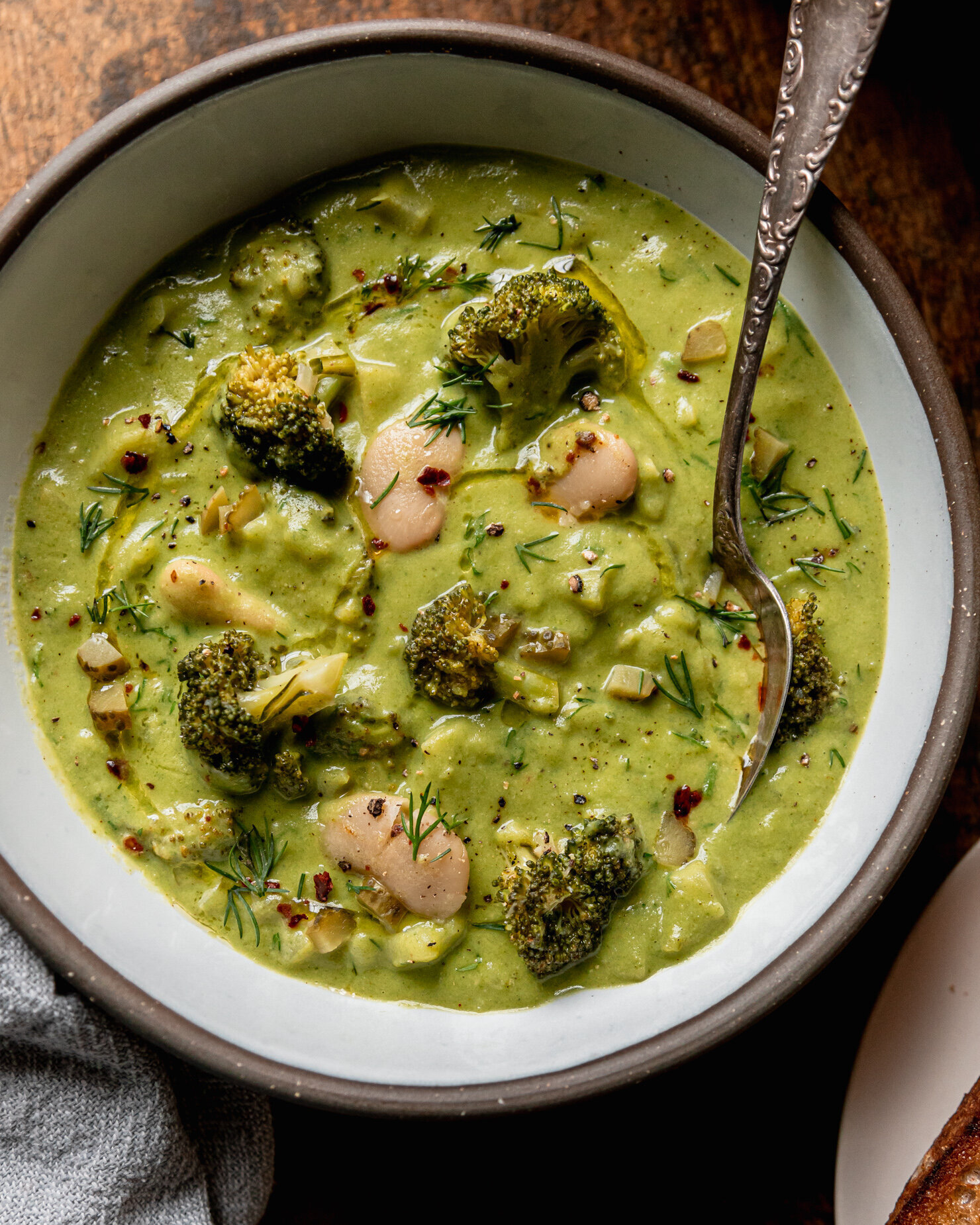 An up close, overhead shot shows a bowl of creamy, light green broccoli dill pickle soup with white beans and potatoes. A spoon is sticking out of the bowl and the soup is garnished with chili flakes, chopped fresh dill, and swoops of olive oil.