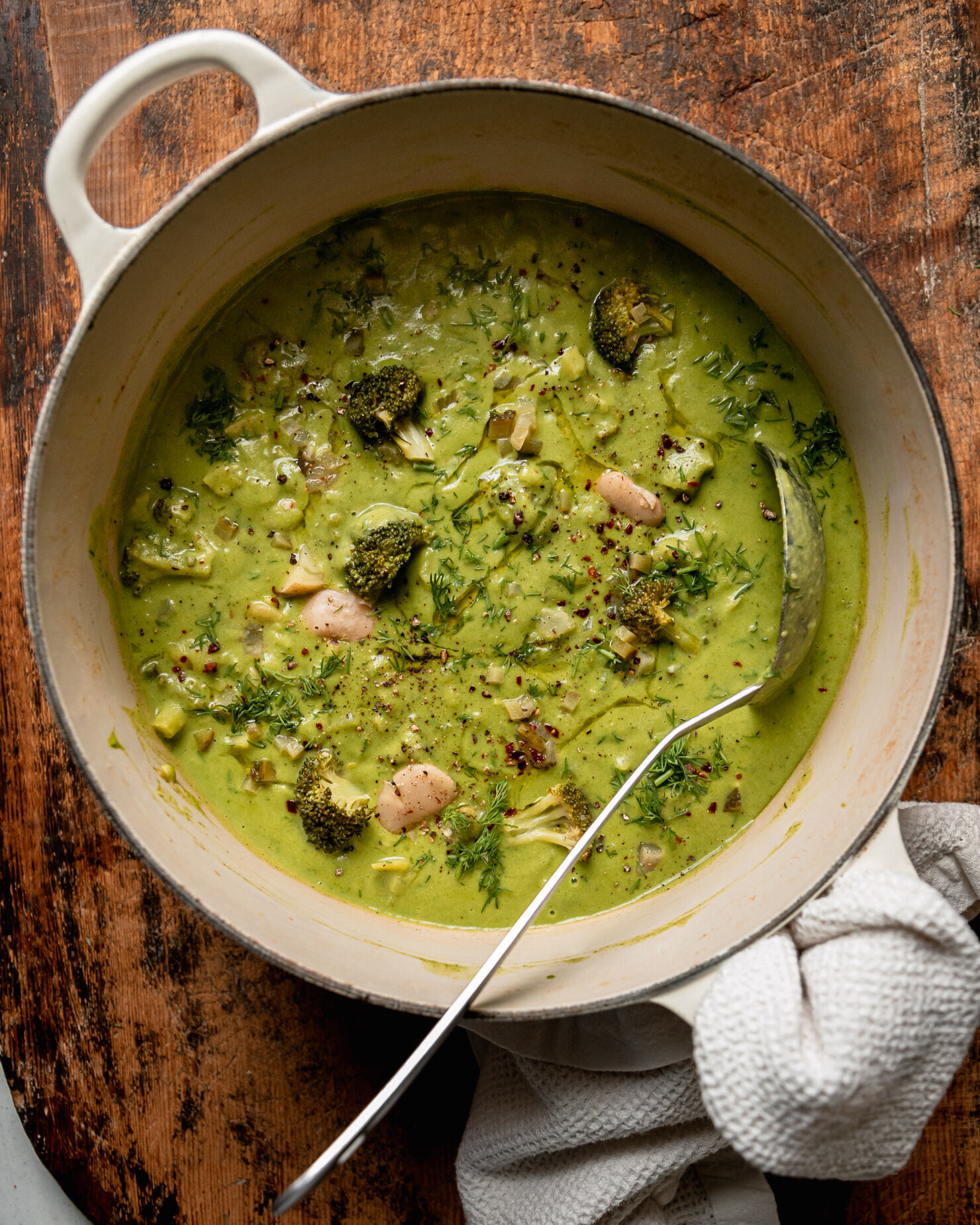 An overhead shot shows a pot of broccoli dill pickle soup with white beans and potatoes. The soup is creamy and light green in colour with a ladle sticking out of the pot.