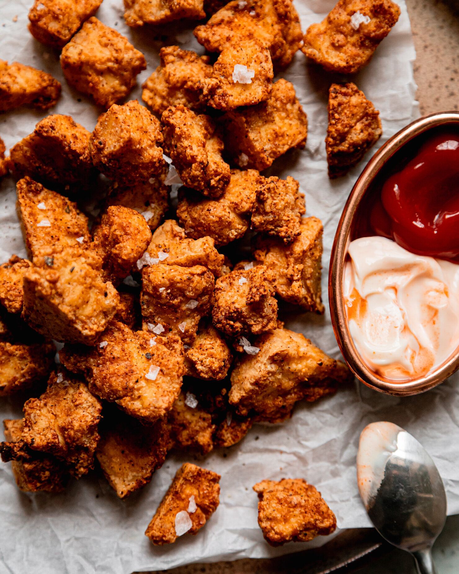 An overhead shot shows crispy torn pieces of tofu on a parchment-lined plate. A carafe of ketchup and spicy mayo for dipping is seen to the side.