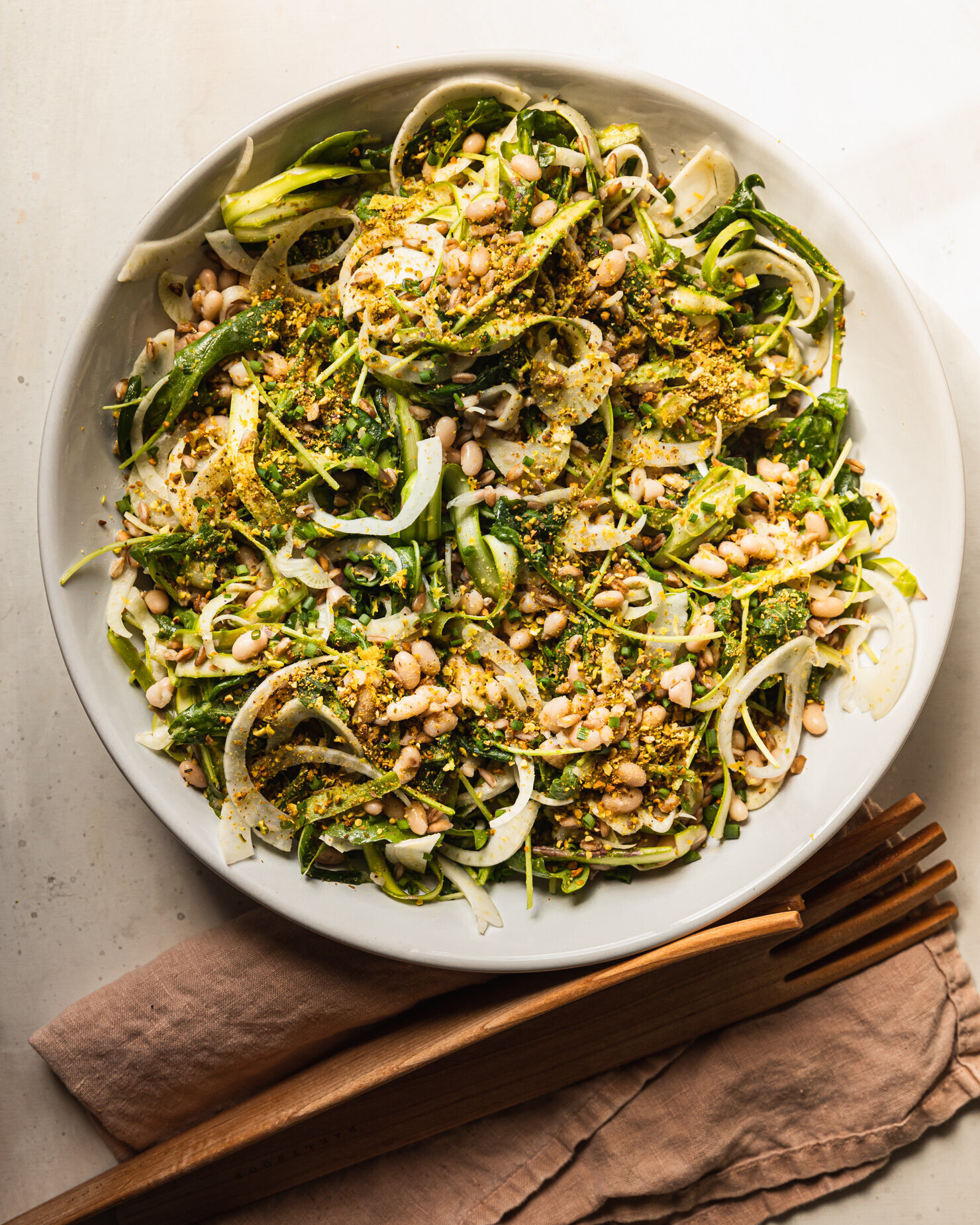 An overhead shot shows a wide bowl filled with lemony shaved fennel and asparagus salad with cooked farro, white beans, and finely chopped pistachios.