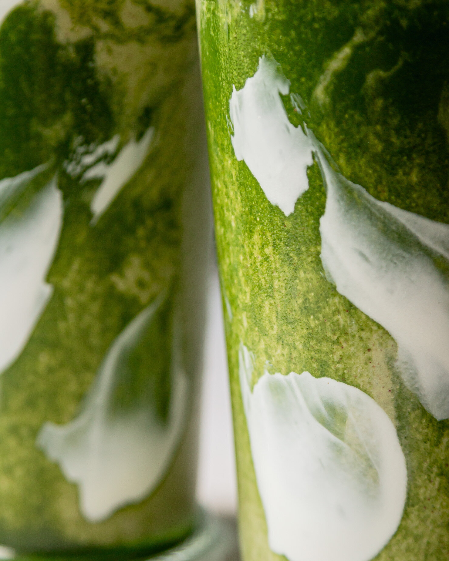 An up close shot shows two glasses of matcha smoothie almost layered on each other. Through the glass you can see swirls of light green, dark green and white.