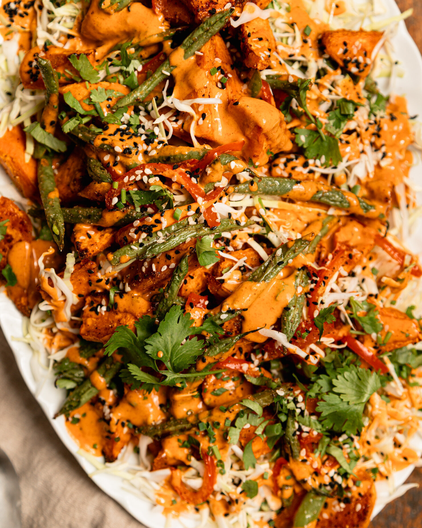 An up close, overhead shot shows a Gochujang peanut sweet potato salad with green beans, red bell peppers, shredded cabbage, and chopped herbs. The roasted vegetables are piled on top of the cabbage on an oval platter and topped with peanut Gochujang dressing and sesame seeds.