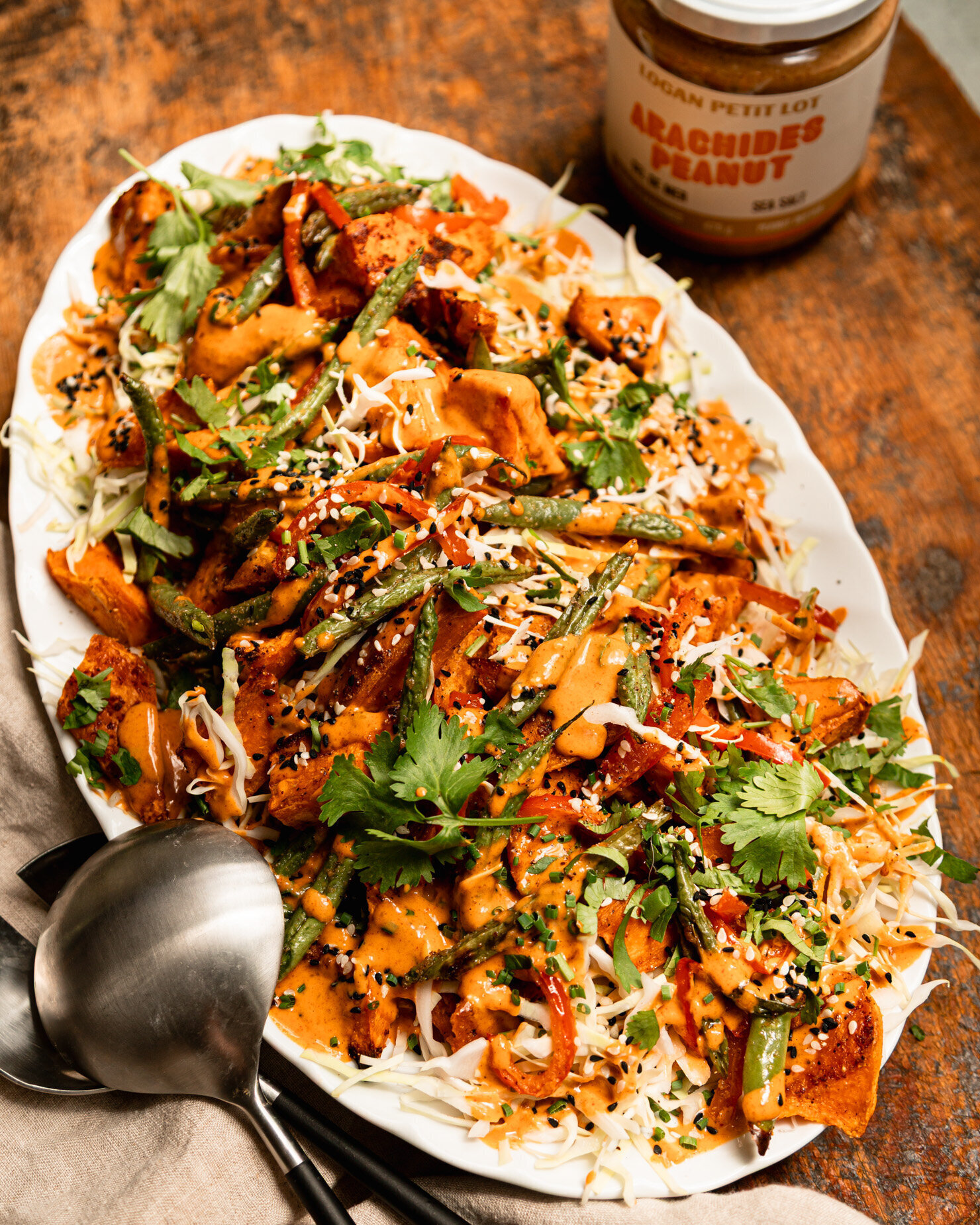 A 3/4 angle shot shows a Gochujang peanut sweet potato salad with green beans, red bell peppers, shredded cabbage, and chopped herbs. The roasted vegetables are piled on top of the cabbage on an oval platter and topped with peanut Gochujang dressing and sesame seeds. A jar of Logan Petit Lot peanut butter and serving tongs are seen nearby.