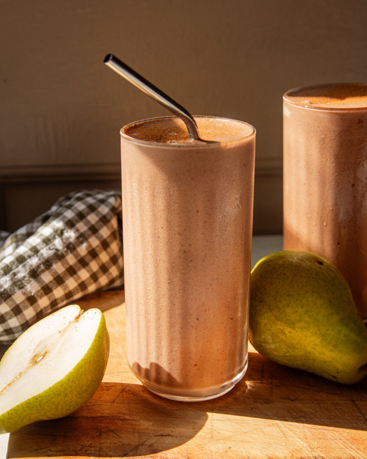 A head-on shot shows a chocolate pear smoothie on a wooden board in direct sun light. A stainless straw sticks out of the smoothie and whole pears are seen nearby.