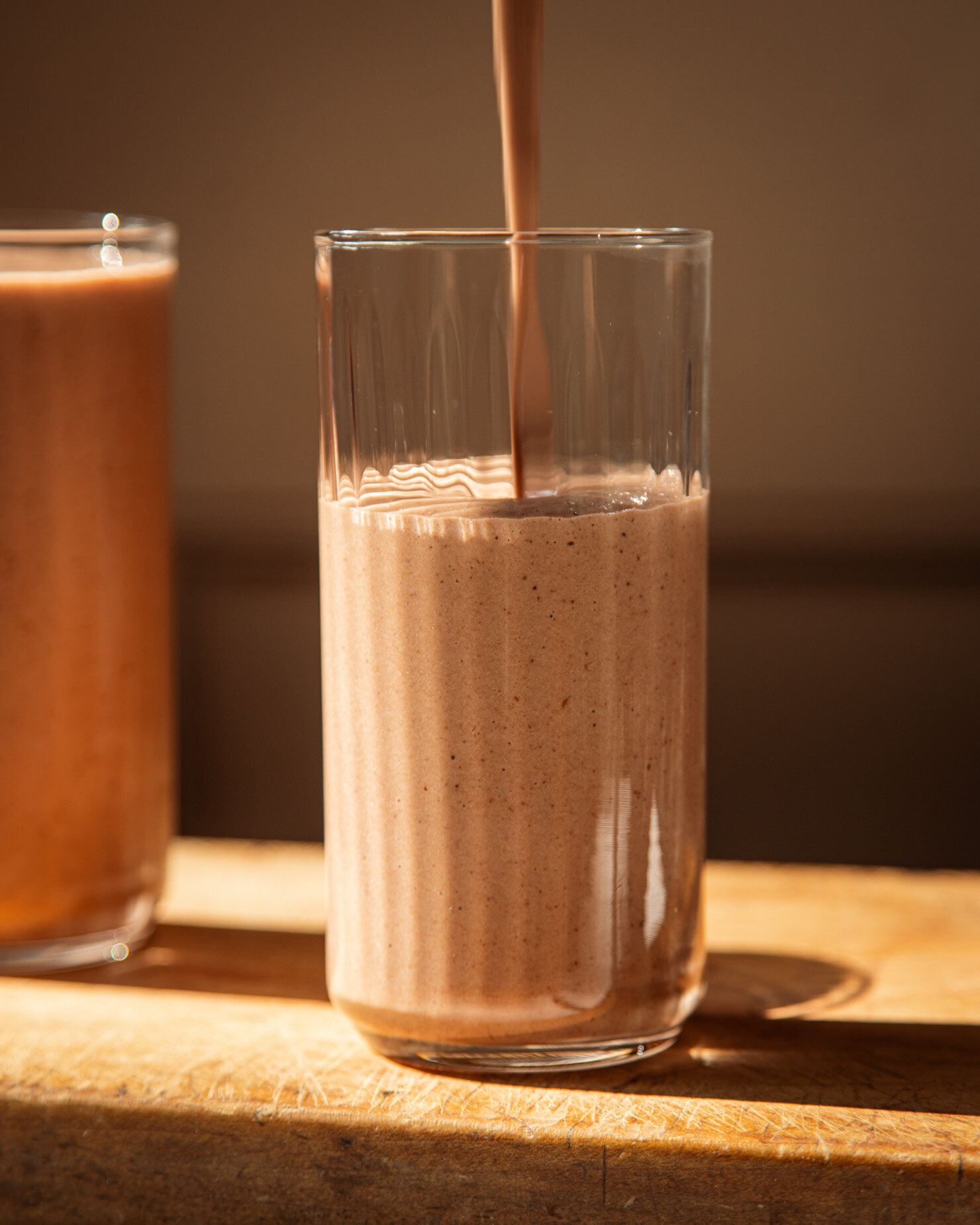 A head-on shot shows a chocolaty smoothie being poured into a glass in bright sun light.