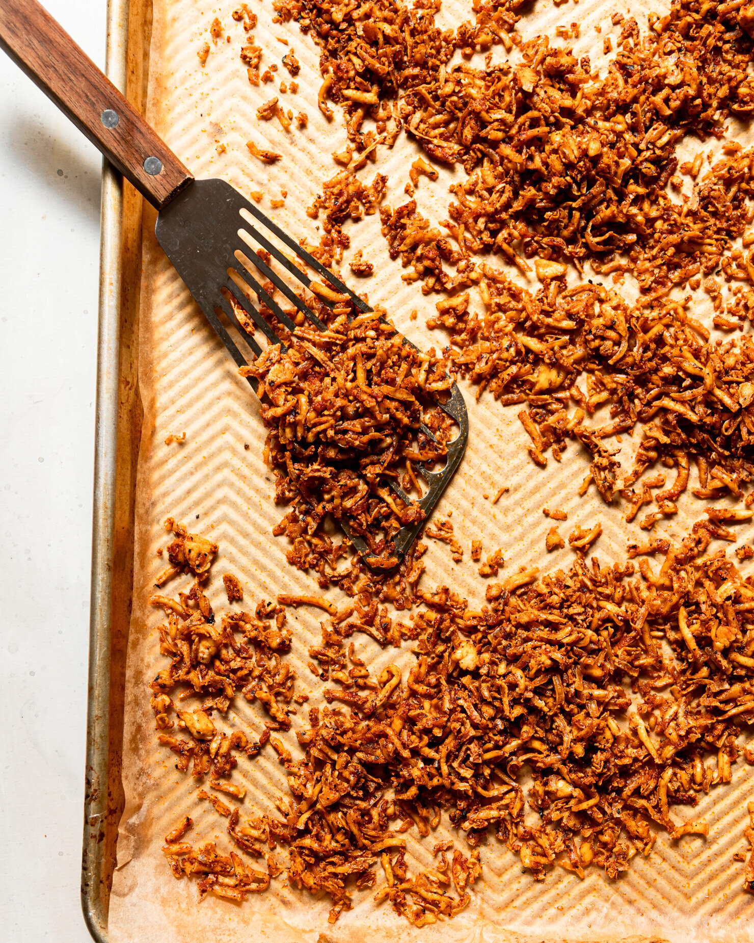 An overhead shot shows a baking sheet filled with crispy and chewy baked shredded tofu. A metal spatula is seen to the side.