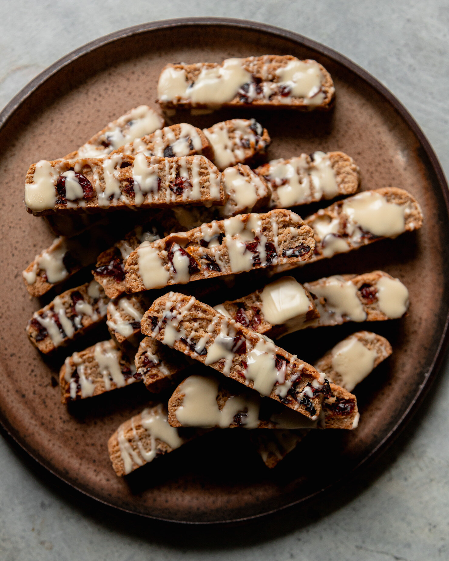 An overhead shot shows a plate piled high with vegan orange cranverry biscotti drizzled with melted vegan white chocolate.