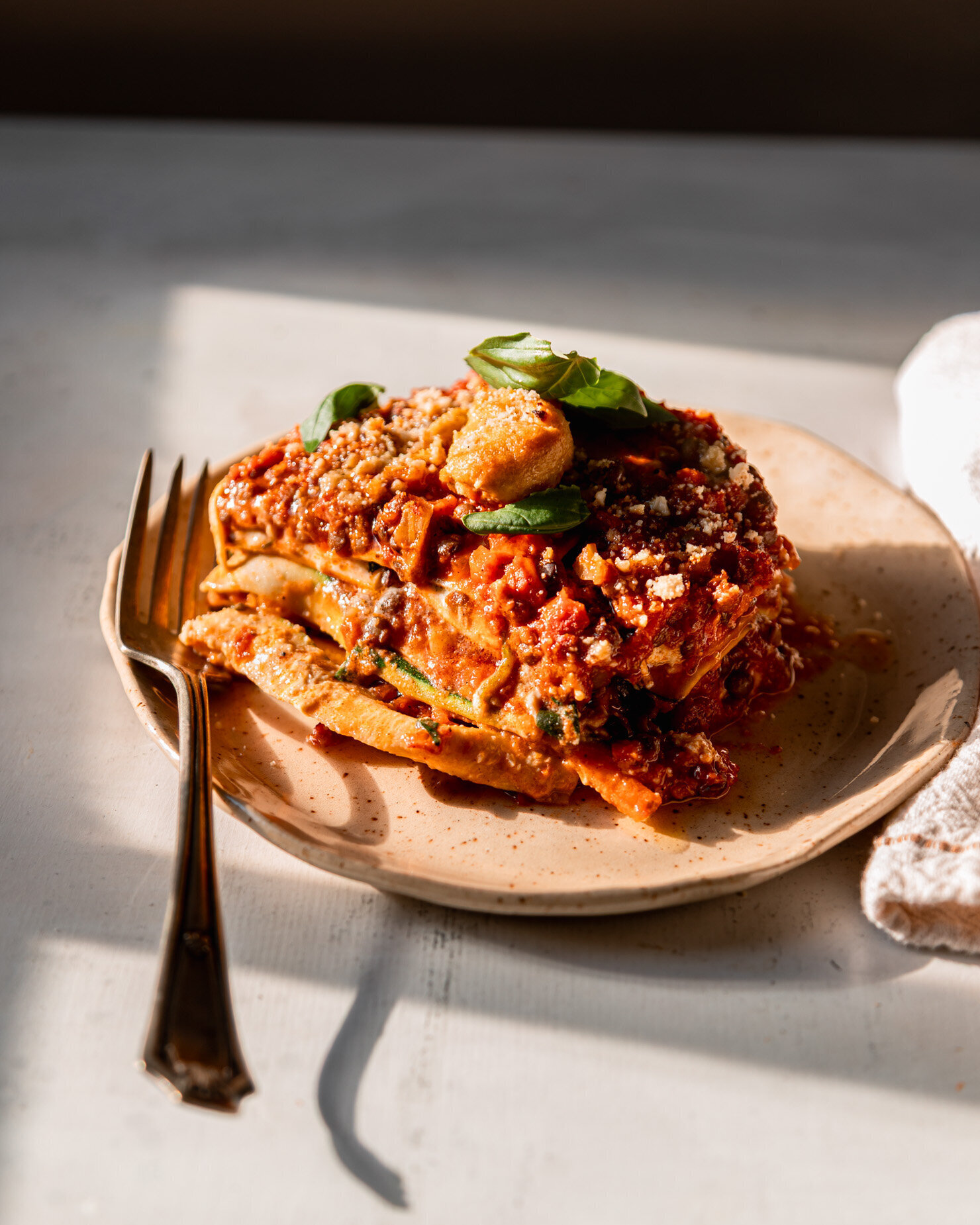 A slight 3/4 angle shows a piece of vegan lasagna on a plate in full sun. A fork is resting alongside on the plate.