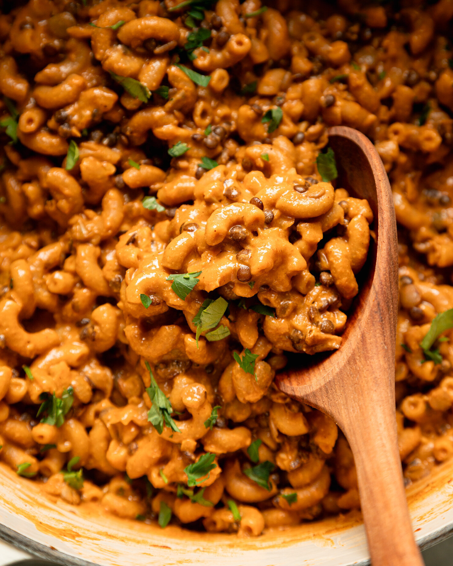 An up close, overhead shot shows a vegan hamburger helper-style mixture with lentils, elbow macaroni, and chopped parsley. A wooden spoon is sticking out of the mixture.