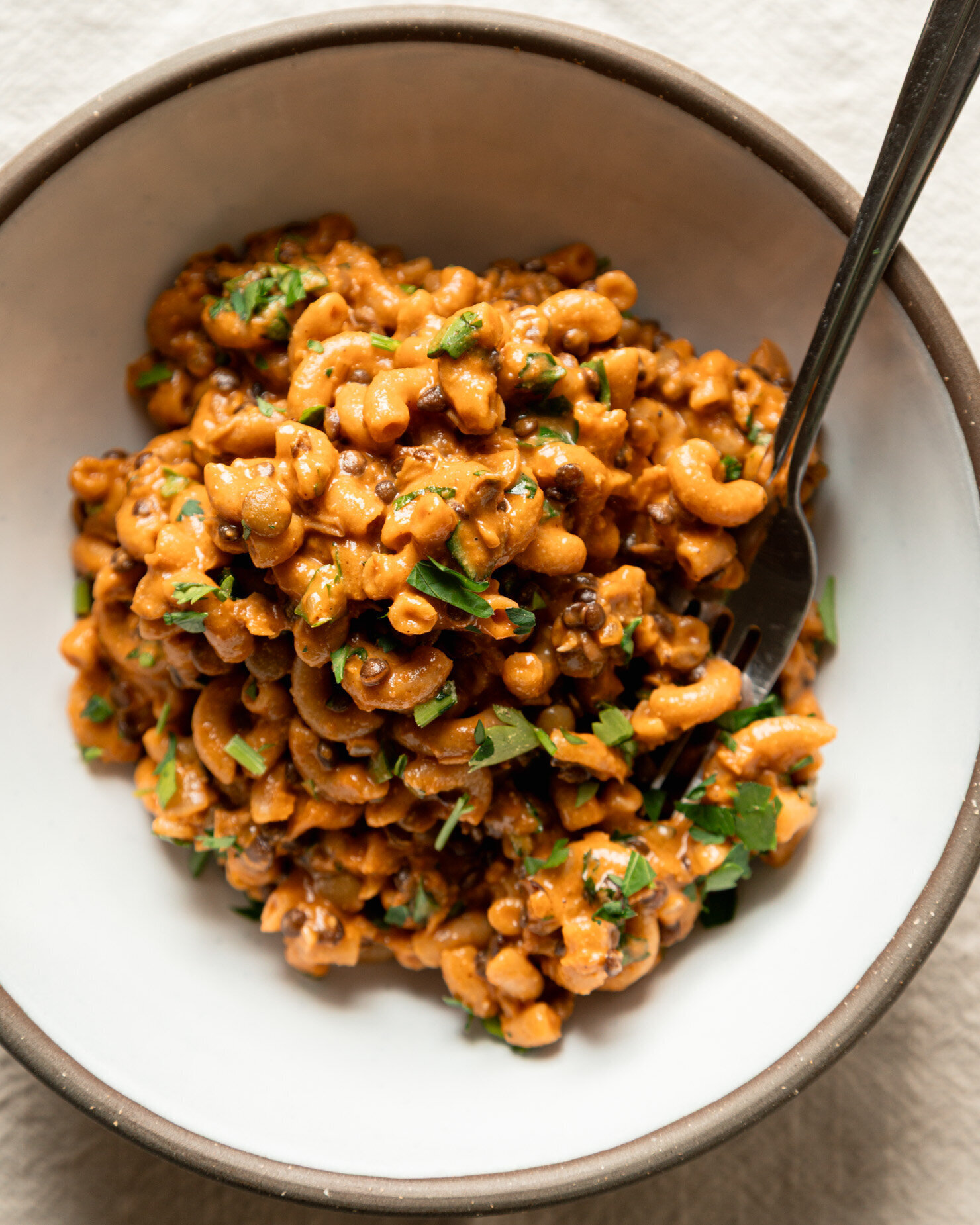 An overhead shot shows an individual serving of vegan hamburger helper with lentils, elbow macaroni, and chopped parsley. A fork is sticking out of the serving in a wide ceramic bowl.