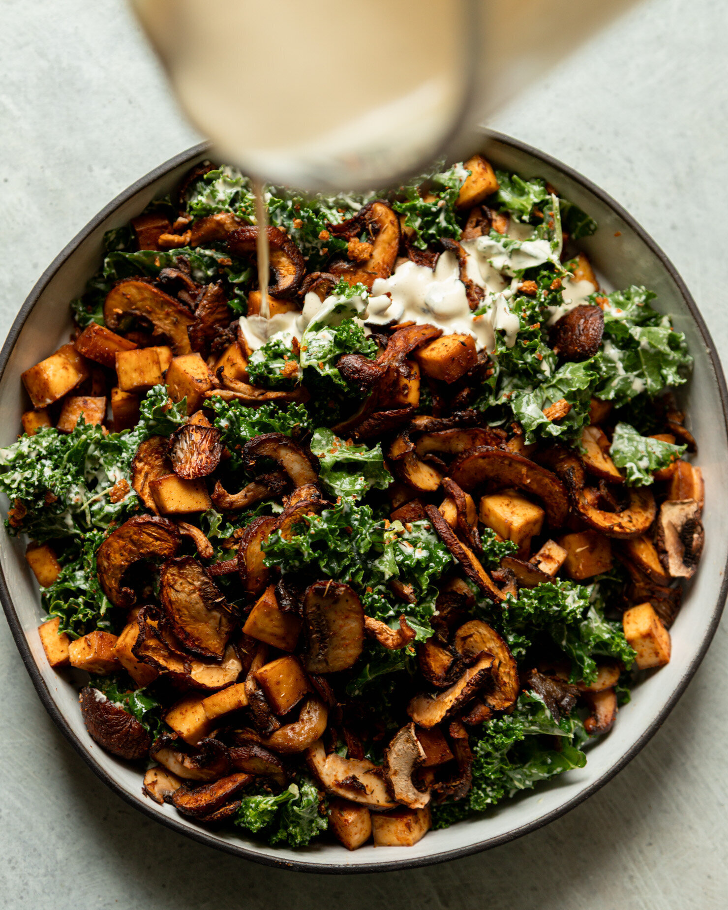 An overhead shot shows creamy garlic cashew dressing being poured over dressed chopped kale, roasted mushrooms, and smoked tofu.