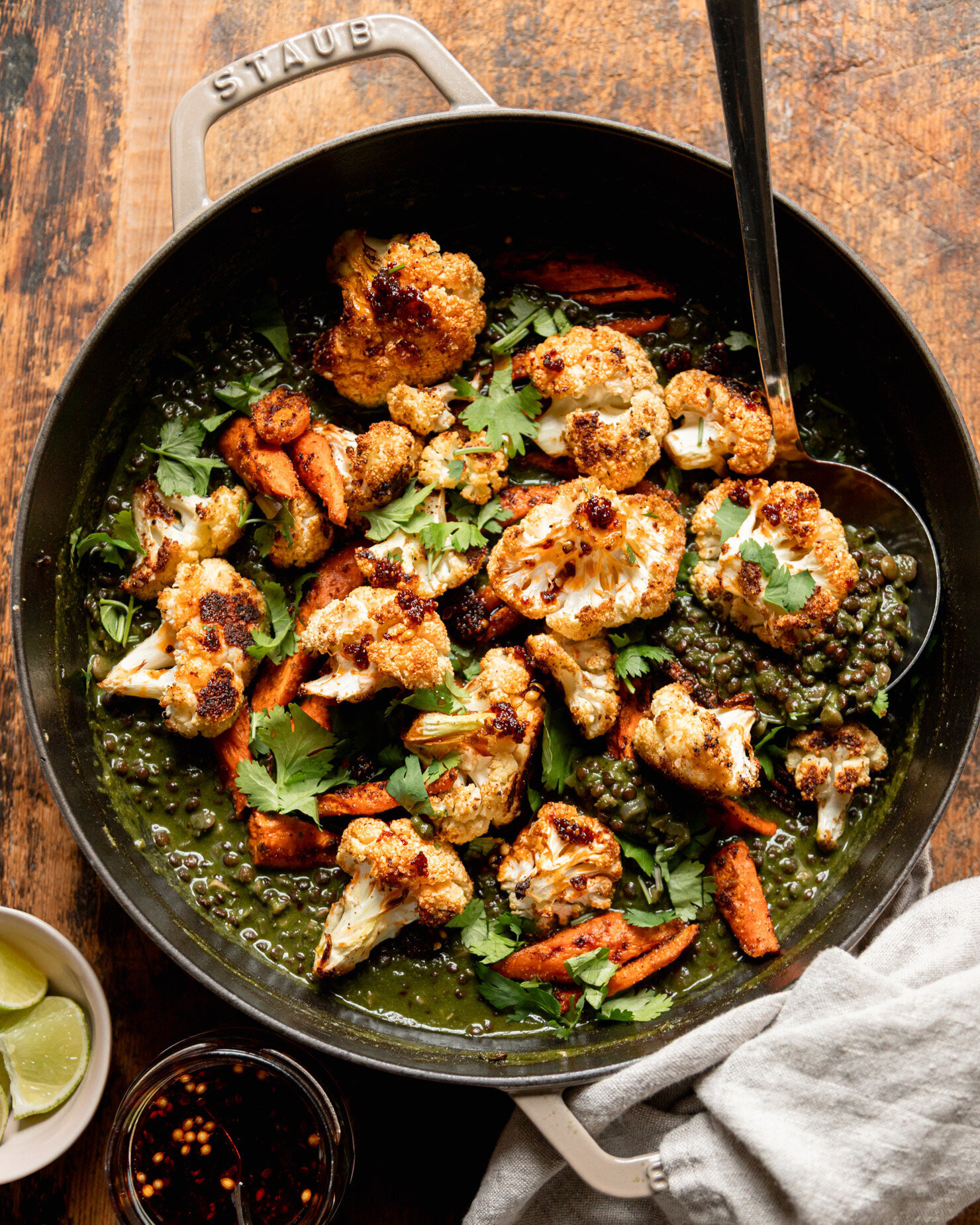 An overhead shot shows a braiser pot filled with spinach and coconut stewed lentils with roasted carrots and cauliflower on top. A ladle is sticking out of the pot. Chopped cilantro garnishes the dish. Lime wedges and chili crisp are seen to the side.