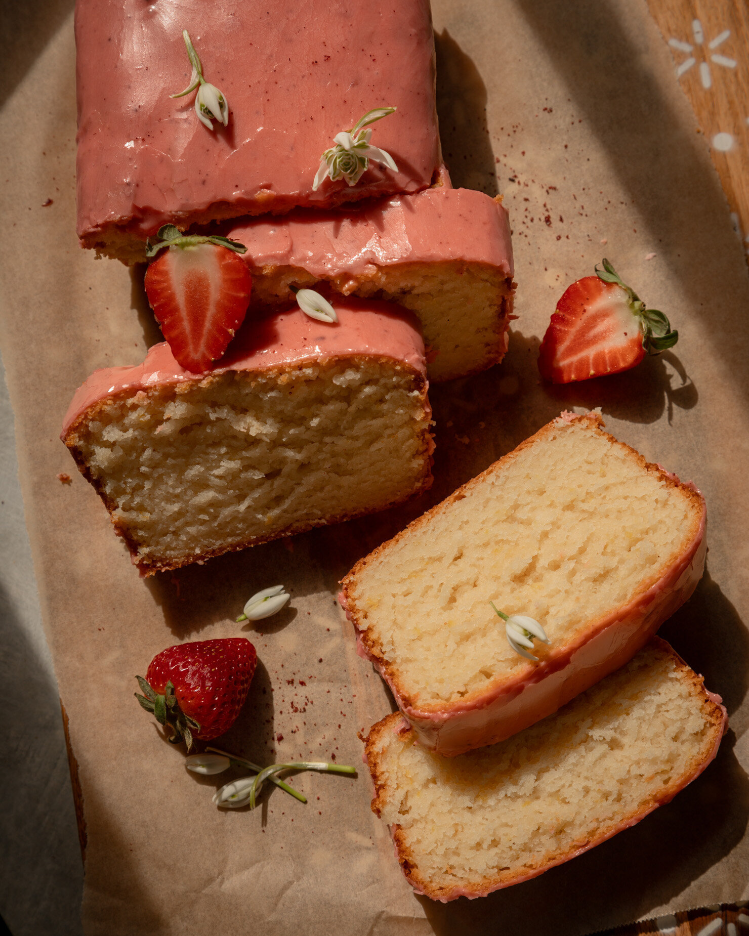 An overhead shot shows a sliced vegan lemon loaf with strawberry sumac glaze. Whole and halved strawberries as well as snow drop flowers are seen nearby. The photo is taken in warm sun light.