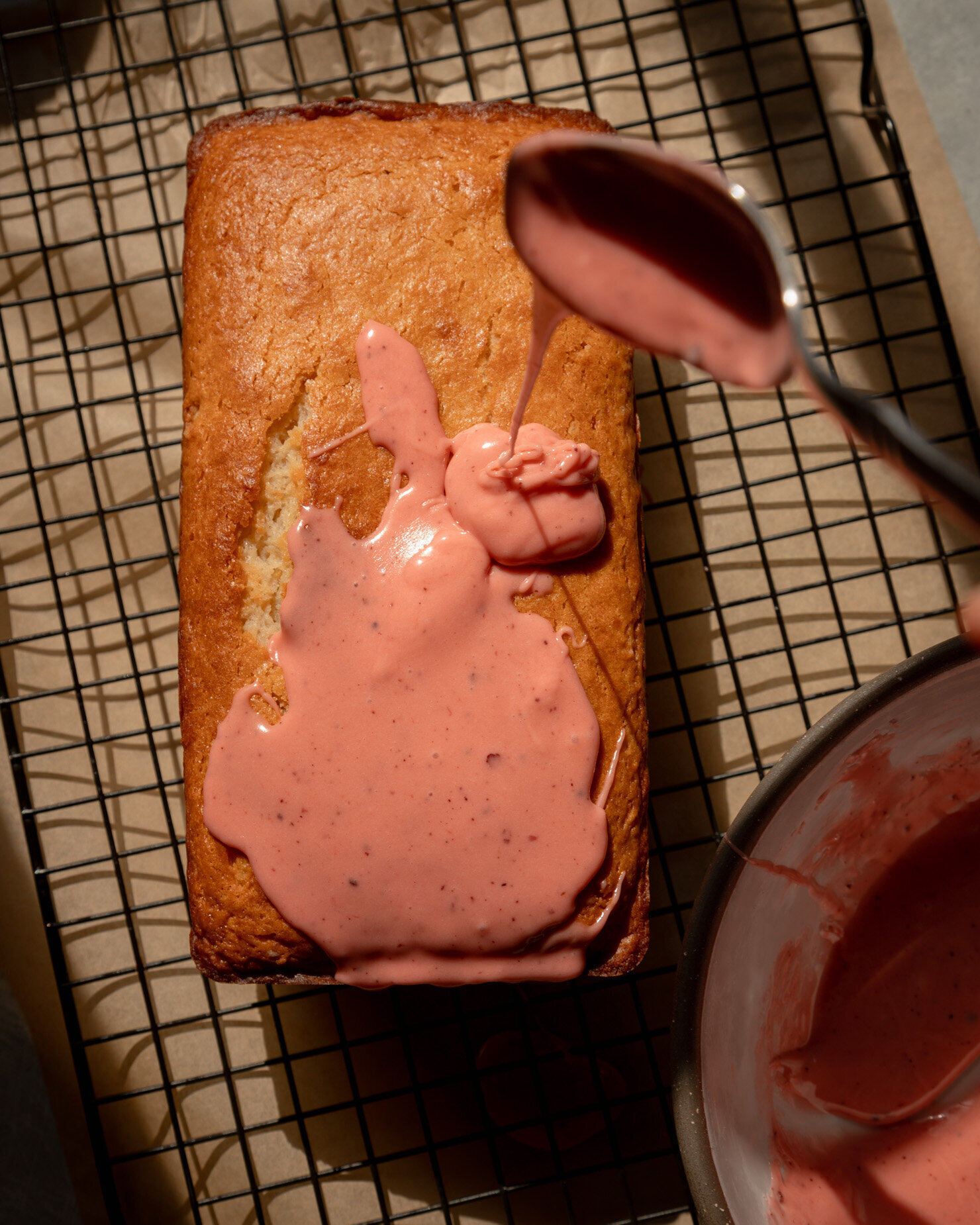 An overhead shot shows a spoon drizzling strawberry sumac glaze over a vegan lemon loaf that is resting on a cooling rack.