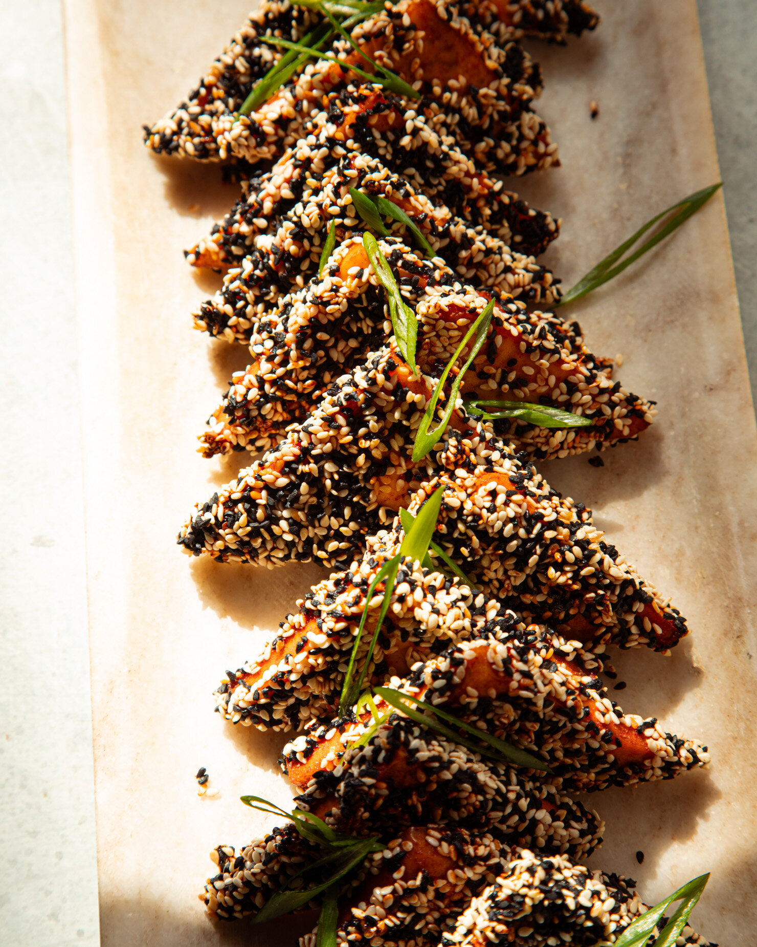 An overhead shot shows a row of sesame-crusted tofu triangles posed upright on a platter in direct light. The tofu is garnished with sliced green onions.