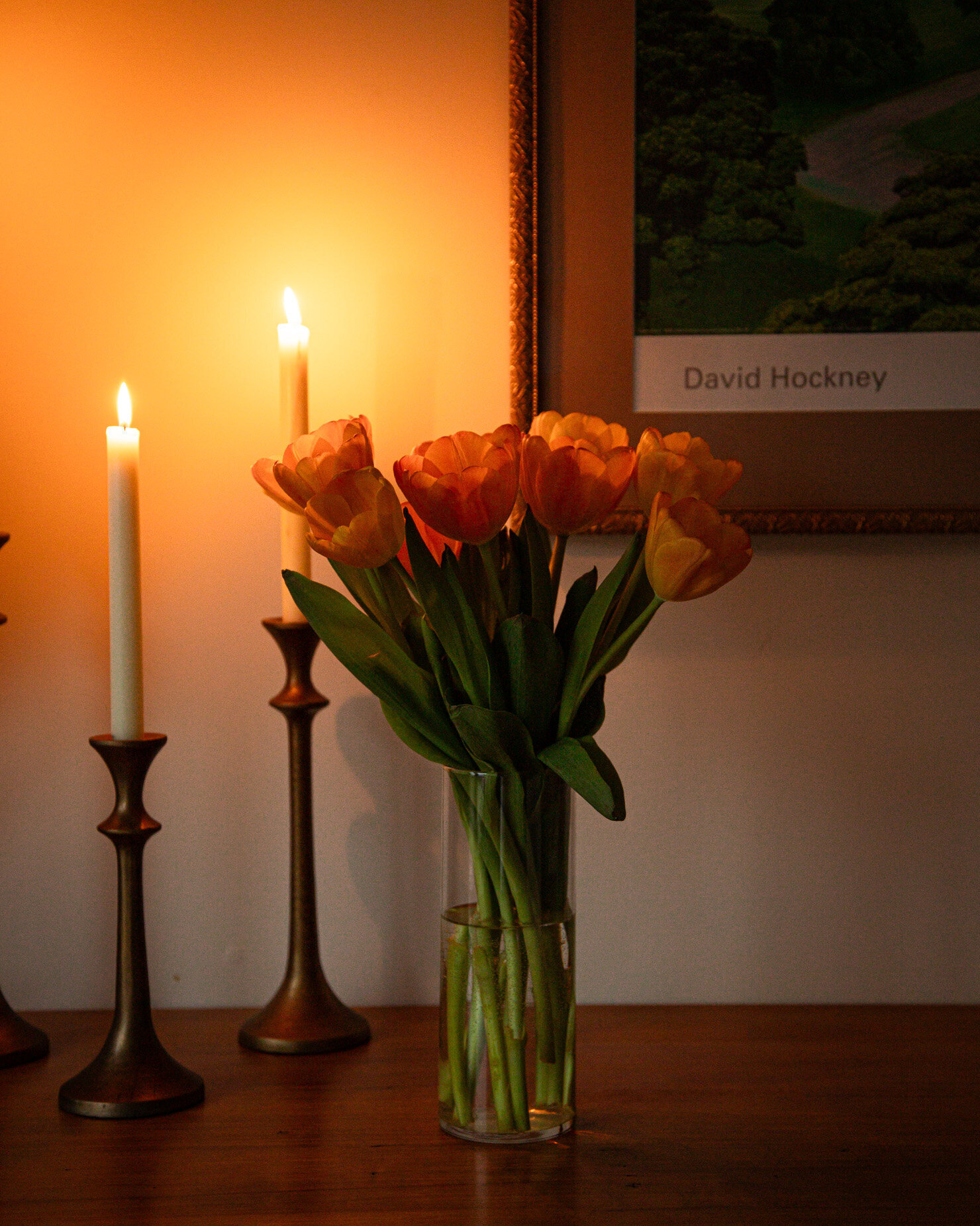 A head-on shot shows a fully bloomed bouquet of tulips in a base in moody lighting. Two lit candles in brass holders nearby are adding a little glow to the scene.