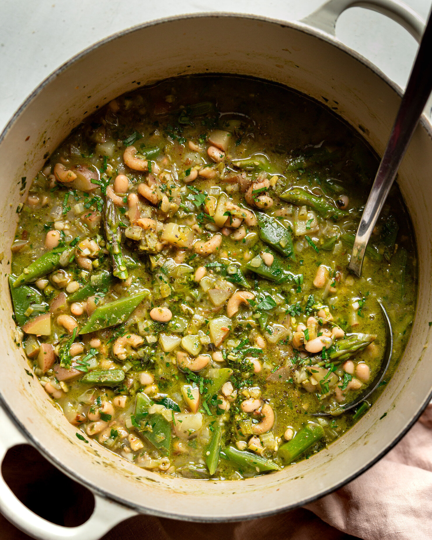 An overhead shot shows a Dutch oven filled with spring green minestrone soup. A ladle is sticking out of the soup to the side.