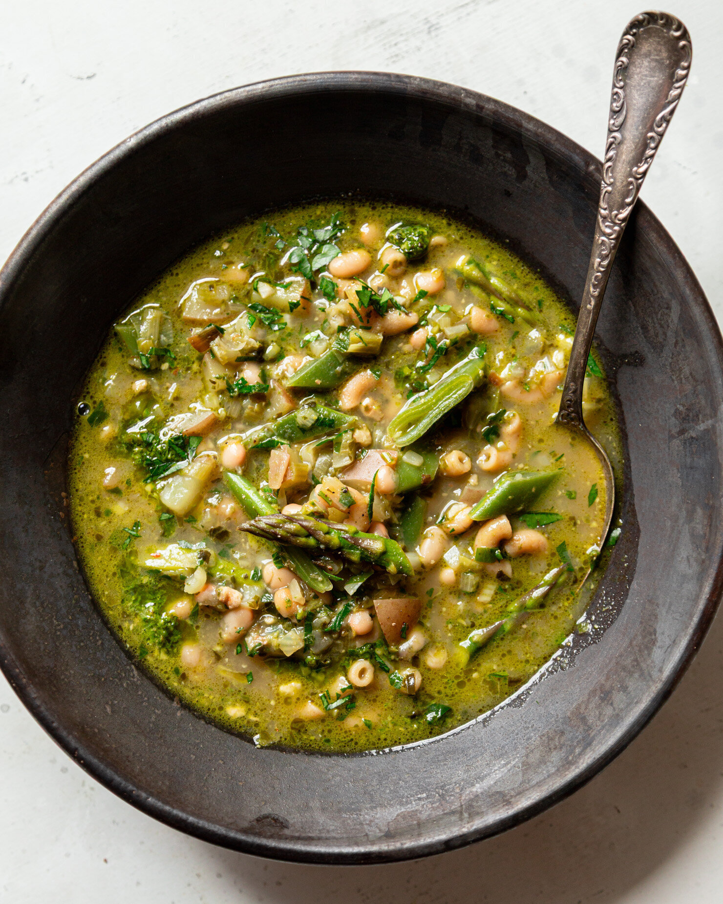 An overhead shot shows an individual serving of spring green minestrone soup in a wide, dark bowl. A spoon is sticking out of the side, and the soup is garnished with ground black pepper and chopped parsley.